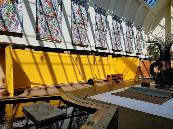 Long bright yellow temporary wall in a high-ceilinged atrium with floral banners above and a reflecting pool in the foreground, Omaha 2023